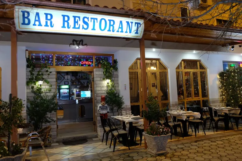 The entrance of Restorant 4 stinët at night: the Bar Restorant sign, the open bar and set tables outside